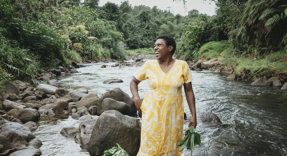 Eine Frau steht lachend in einem flachen Fluss mit vielen Felsbrocken. 
