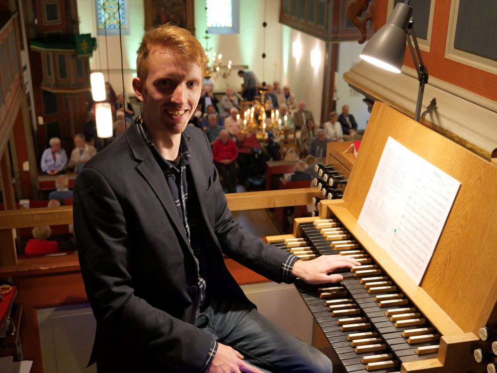 Organist sitzt in der Kirche an der Orgel. Im Hintergrund unten Publikum.