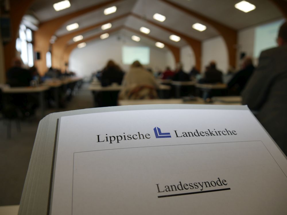 Man sieht im Vordergrund eine Mappe, auf der steht Lippische Landeskirche und Landessynode. Im Hintergrund verschwommen ein Tagungsraum mit Menschen. 